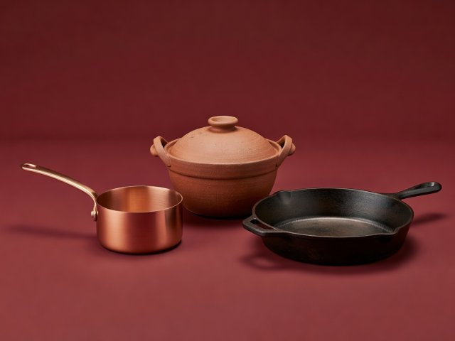 Soft-lit 3D render of an earthenware donabe pot, copper saucepan and seasoned cast-iron skillet on a burgundy background